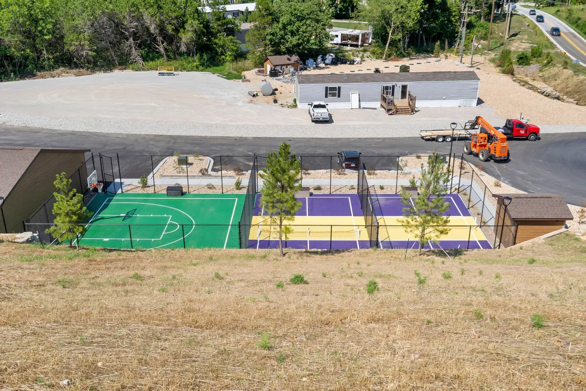 The image shows a vibrant outdoor sports area with a basketball court and tennis courts nestled in lush greenery. Several groups can be seen playing games and enjoying outdoor activities, creating an energetic and communal vibe.