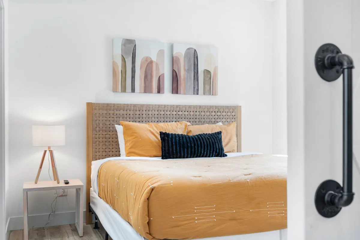 This image features a king bed adorned with a golden orange duvet, decorative pillows, abstract wall art, and a nightstand with a lamp. Soft natural light filters into the room, conveying a sense of comfort and tranquility suitable for restful nights.