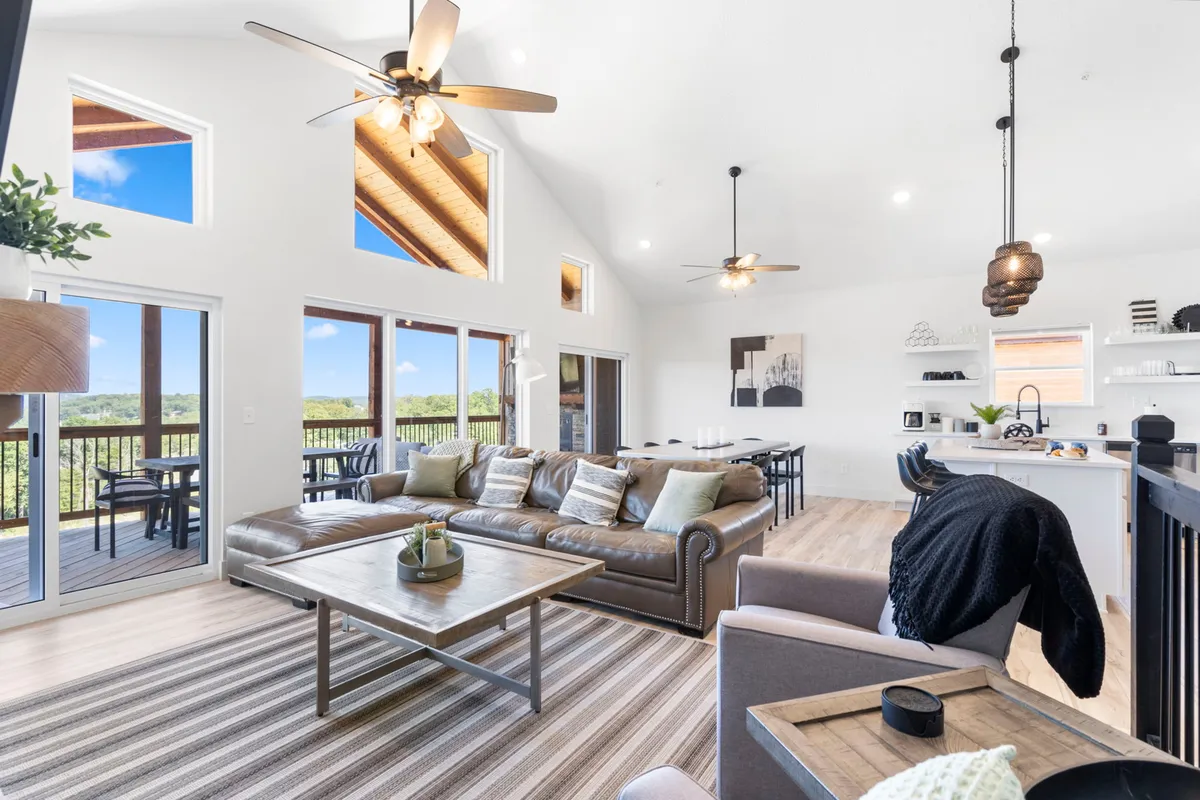 This interior scene features a large leather sofa and wooden coffee table set against panoramic windows that offer a scenic lake view. The space is decorated with stylish artwork and is ideal for relaxing or entertaining, with an emphasis on comfort and light.