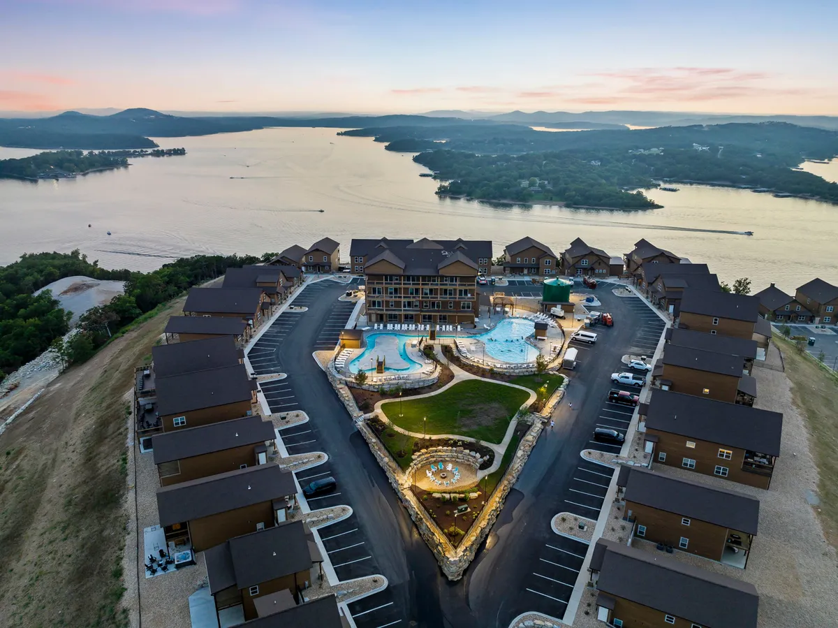 A lively exterior scene featuring a large swimming pool, outdoor seating area, and a green lawn with a stunning view of the lake and surrounding mountains. People are visible swimming and sunbathing, creating a playful, social, and relaxed mood that mirrors the lakefront fun and group activities mentioned in the text.