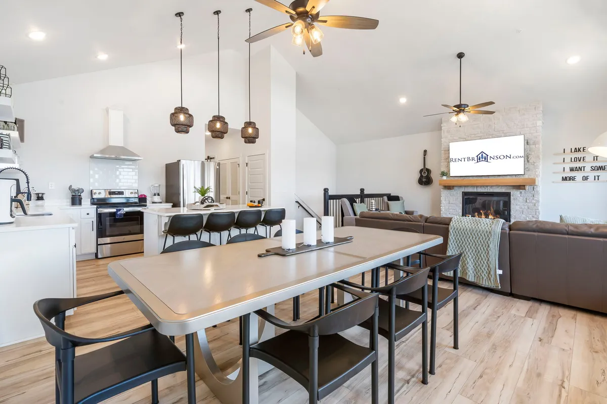 The image presents an inviting view of the dining table, kitchen island, fireplace, and a television in an open layout. Large windows and a contemporary chandelier fill the space with brightness, while the arrangement encourages group meals and togetherness.