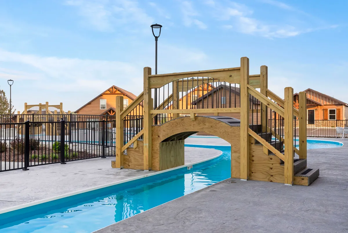 An inviting exterior scene featuring a wooden bridge over a swimming pool, surrounded by buildings and children playing in the water. The sunset sky adds a warm, relaxed atmosphere, evoking the sense of outdoor enjoyment and family-friendly activities.