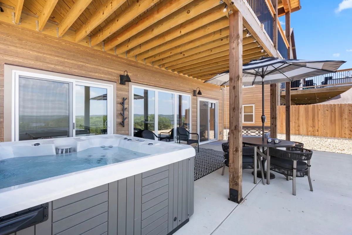 This exterior image showcases the main deck area with a hot tub, outdoor dining table, large glass windows, and wooden beams. People are seen relaxing in the hot tub and dining outdoors while enjoying the expansive lake views.