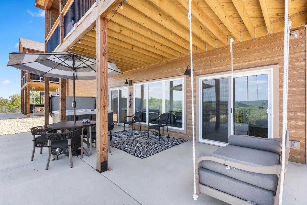 A stylish exterior dining area with a table set for outdoor meals, swing chairs for relaxation, an umbrella for shade, and large sliding glass doors that connect the indoor and outdoor spaces. The area is designed for both casual gatherings and special celebrations, with hints of twilight ambiance.