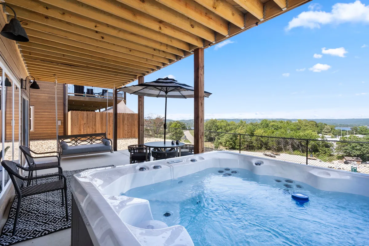 The image showcases the exterior of the home, with a luxurious hot tub positioned to take in sweeping lake views. Comfortable outdoor seating invites guests to relax, while the terrace provides space for morning coffee or evening gatherings, all set against a tranquil sunset backdrop.